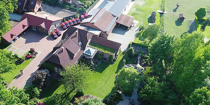 Eine Luftaufnahme eines großen, gepflegten Anwesens mit roten Dächern und einem weitläufigen Garten voller Bäume und Blumen.