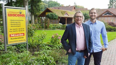 Zwei Männer stehen lächelnd vor einem Geschäftsschild, umgeben von einem gepflegten Garten mit bunten Blumen.